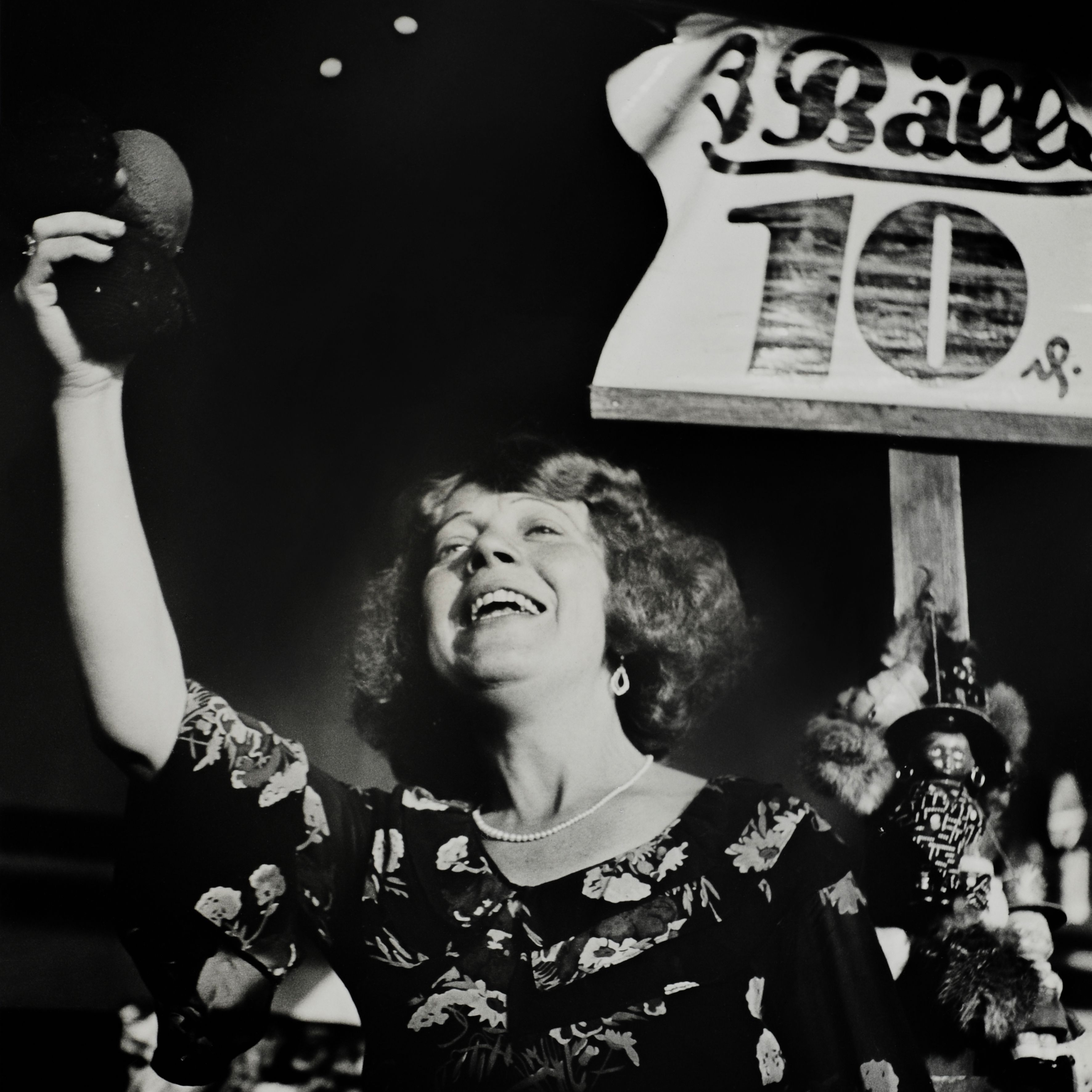 Woman photographed from below. On the top right a placard with "3 balls 10" written on it. Right hand is stretched up. 