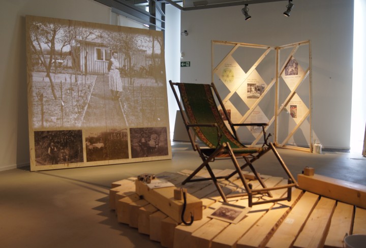 Blick in die Ausstellung: ein Liegestuhl auf einer Holzkonstruktion und eine Leinwand mit historischen Fotos