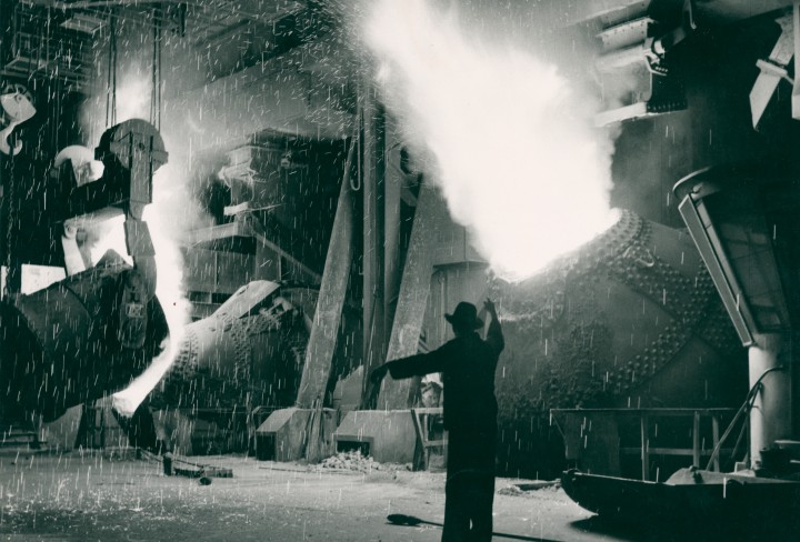Schwarz-weiß-Foto eines Stahlwerks mit Funkenflug und einem gestikulierenden Mann