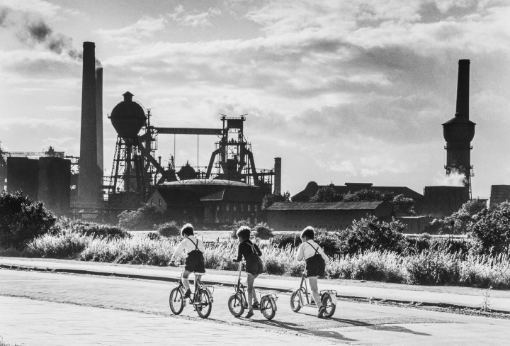 Schwarz/Weiss Aufnahme mit 3 Jungen auf einem Fahhrad vor einer Industrieanlage