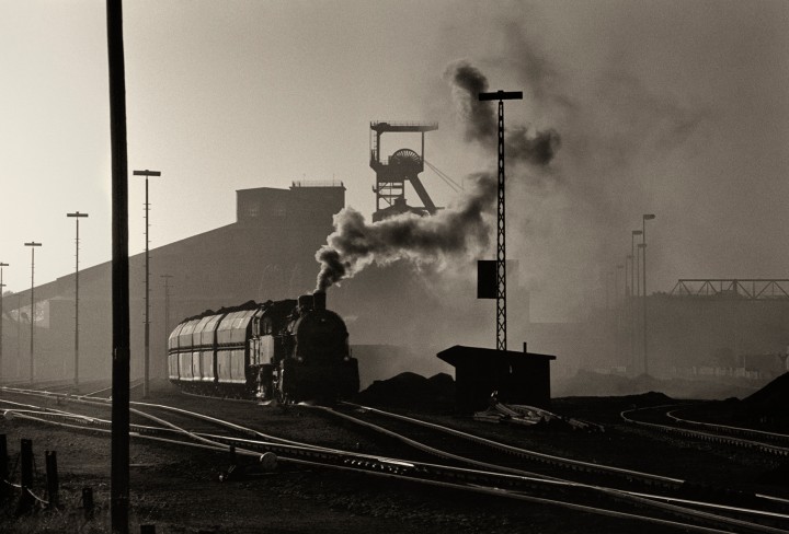 Schwarz-Weiß-Fotografie einer fahrenden Lok vor industriellem Hintergrund
