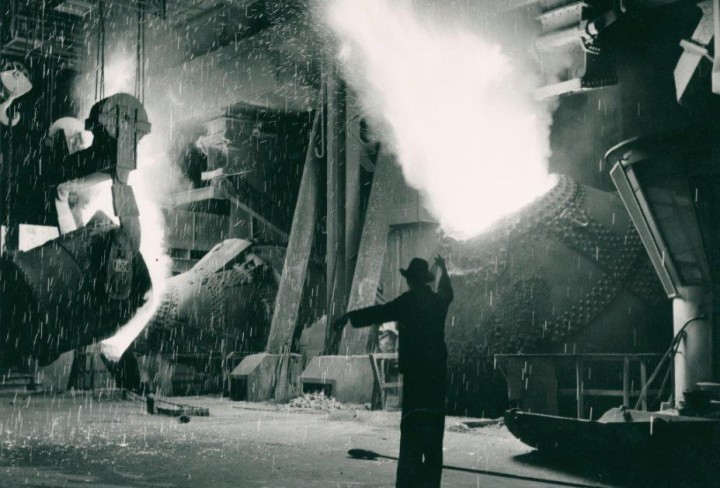 Schwarz-weiß-Foto eines Mannes in einem Werk, dahinter sprühen Funken