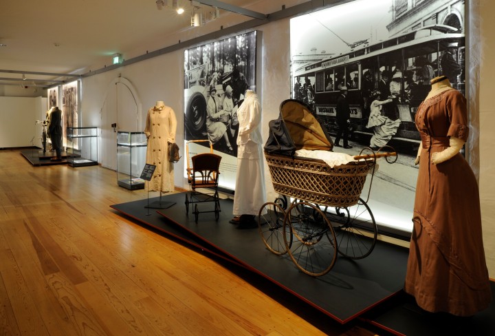 Blick in die Ausstellung mit verschiedenen Kleidungsstücken und einem Kinderwagen auf Podesten