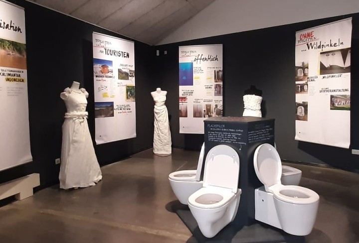Blick in die Ausstellung mit mehreren Texttafeln, Toiletten und Papierkleidern