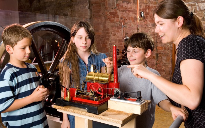 Two boys and a girl look at a model of a steam engine with a woman