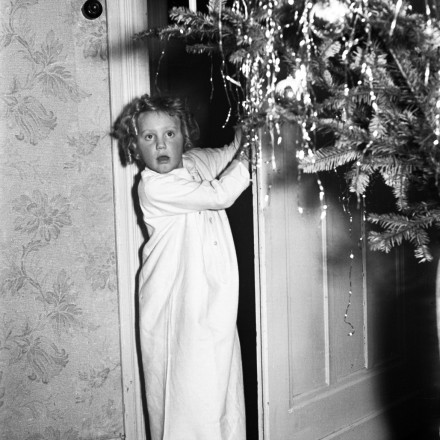 Schwarzweiß-Foto eines Mädchens, das im Nachthemd eine Tür öffnet zu einem Raum, in dem ein Weihnachtsbaum steht
