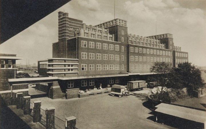 Black and white photograph of a large brick building. There are several cargo trailers and trees in front of the building.