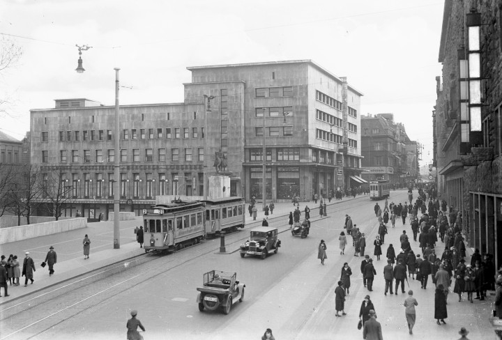 Schwarzweiß-Fotografie einer Straße in der Essener Innstadt. Auf der Straße fährt eine historische Straßenbahn und zwei Autos, auf den Gehwegen befinden sich viele Menschen. Im Hintergrund sieht man ein großes Gebäude.