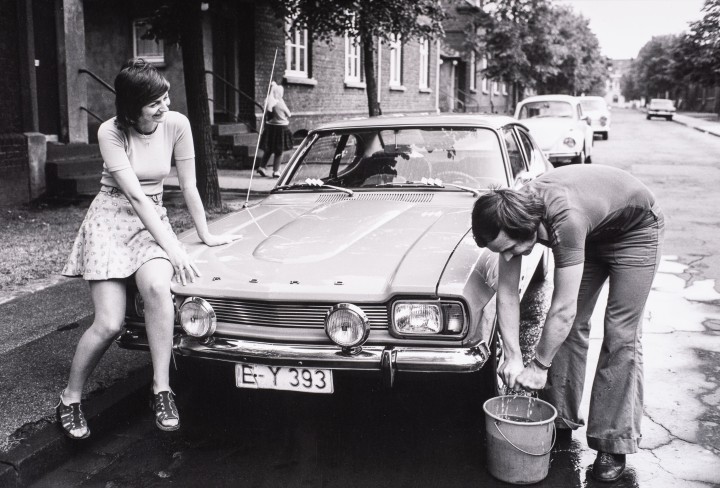 Eine Schwarzweiß-Fotografie aus den 1970er Jahren zeigt einen Mann und eine Frau, die ein Auto waschen