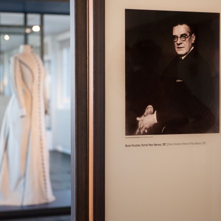 Ausstellungstafel mit einem Porträt von Peter Behrens und ein Hochzeitskleid in einer Vitrine im Hintergrund