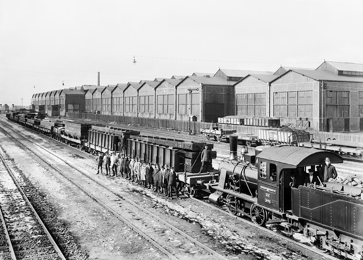 Historisches Schwarzweiß-Foto: Männer stehen nebeneinander vor einem Zug mit Bauteilen auf den Waagons. Im Hintergrund sind große Lagerhallen zu sehen. 