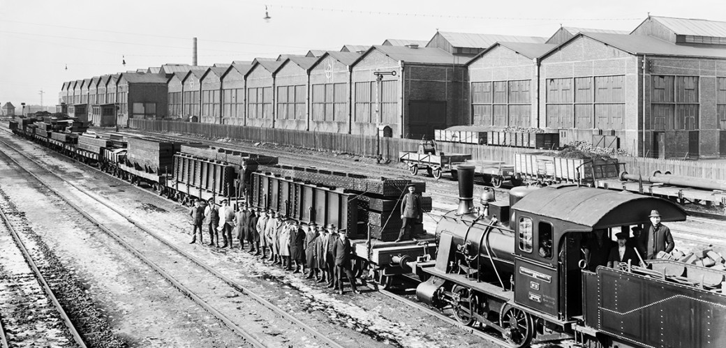 Historisches Schwarzweiß-Foto: Männer stehen nebeneinander vor einem Zug mit Bauteilen auf den Waagons. Im Hintergrund sind große Lagerhallen zu sehen. 