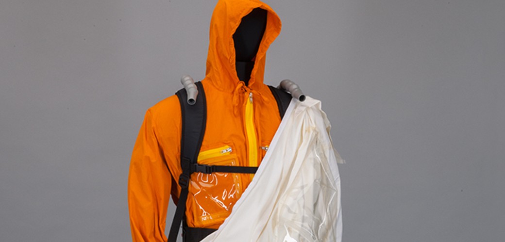 Eine Figurine trägt einen zweiteiligen Arbeitsschutzanzug: Über den inneren, orangenen Anzug ist zur Hälfte ein weißer Schutzanzug gestülpt. Die Puppe trägt dazu türkise Schutzhandschuhe und rote Schuhe