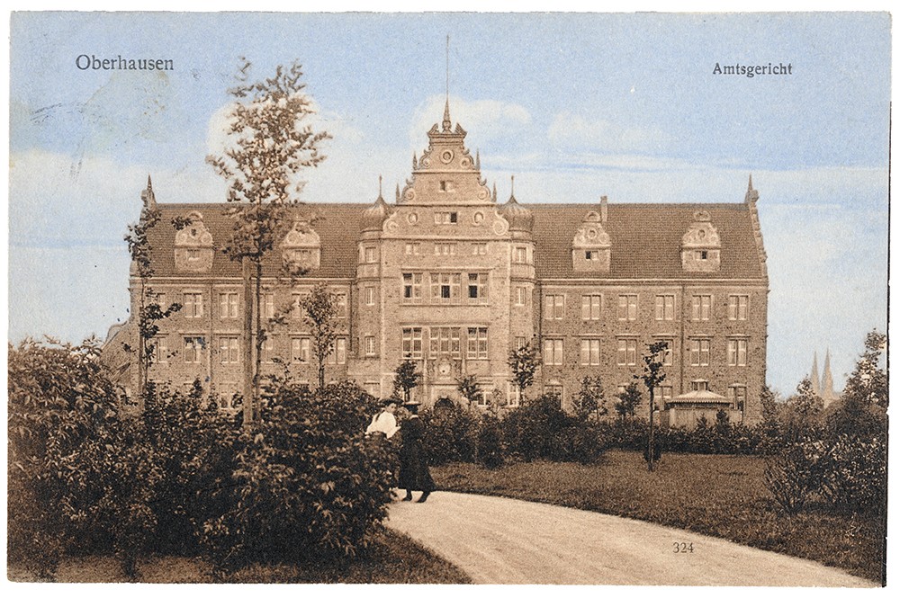 Vorderseite einer Postkarte zeigt ein großes Gebäude, einen Park und zwei spazierende Damen