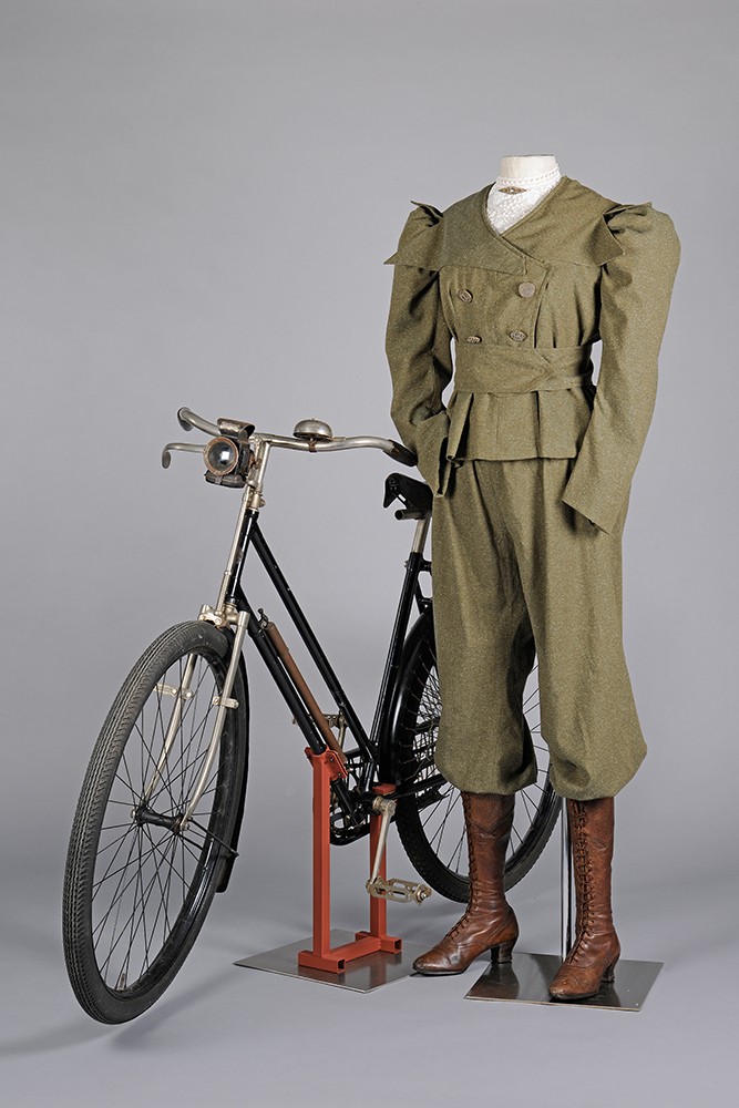 Khakifarbenes Kostüm einer Radfahrerin mit Pluderhose und braunen Lederstiefeln vor einem historischen Fahrrad