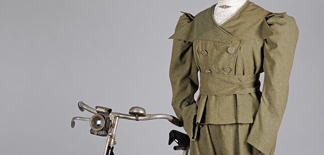 Khakifarbenes Kostüm einer Radfahrerin mit Pluderhose und braunen Lederstiefeln vor einem historischen Fahrrad