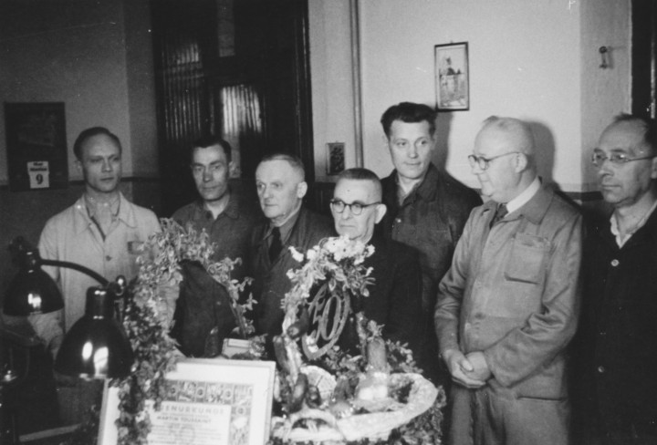 Schwarz-Weiß Foto mit Männern die um einen mit dem Jubilar Tisch stehen
