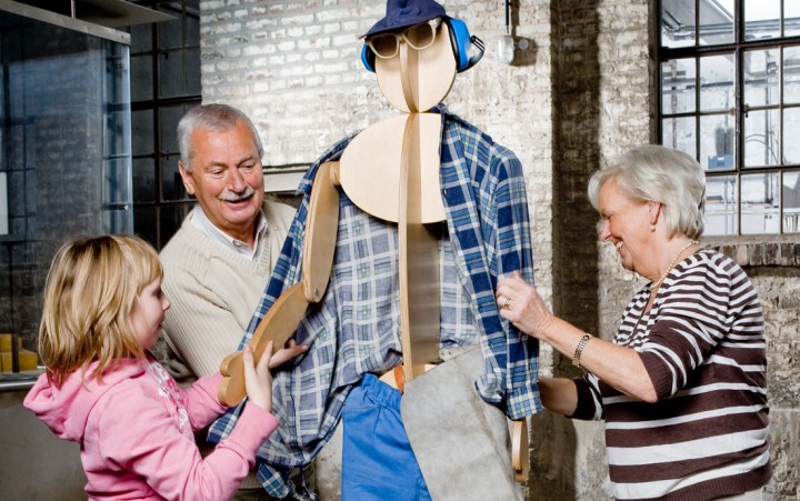 Enkel mit Vater- und Großmutter bekleiden eine Holzkpuppe mit Arbeitskleidung