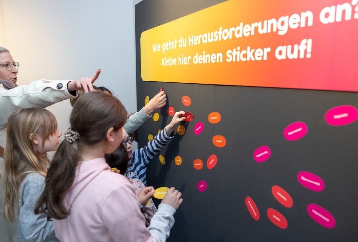 Eine Frau steht mit einigen Kindern vor einer Tafel mit Sticker
