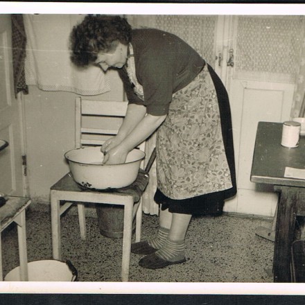 historisches Foto in Schwarz Weiss, auf welchem eine Frau in einer Küche steht und in einer Schüssel etwas mit Wasser spült.