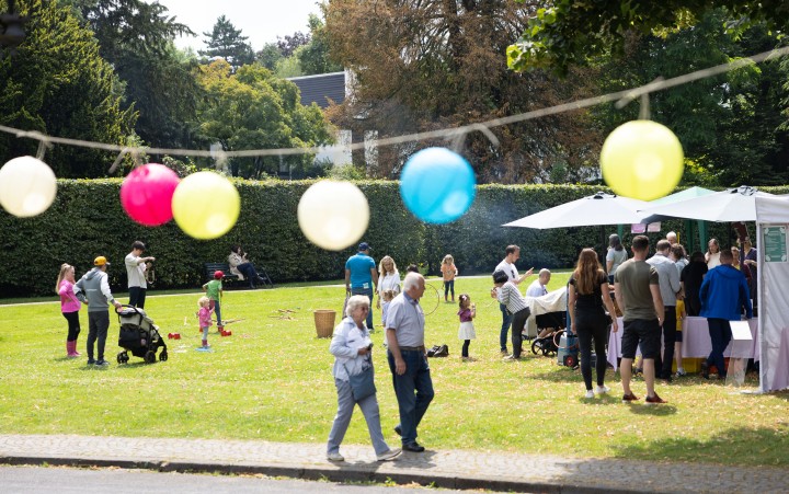 Im Vordergrund gehen zwei ältere Personen auf einem Gehweg entlang und es ist eine bunte Girlande mit Kugeln in Weiß, Pink, Gelb und Blau zu sehen. Im Hintergrund sind Menschen auf einer Wiese. Kinder spielen und Erwachsene stehen an Pavillons. Hinter der Wiese sind hohe Bäume und eine Hecke.