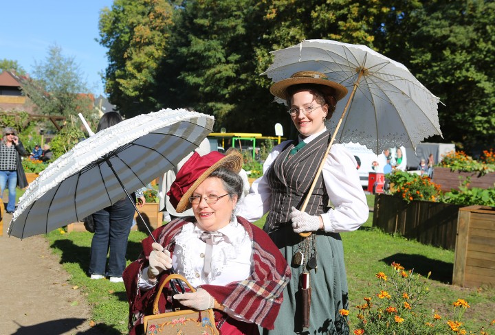 Zwei historisch gekleidete Frauen mit Sonnenschirmen vor einem Garten