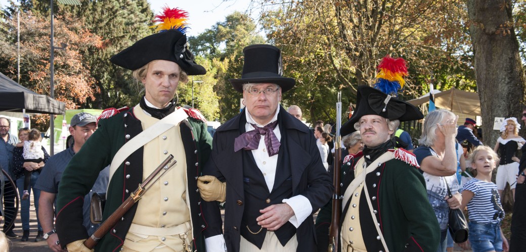 Drei Männer in historischen Kostümen, im Hintergrund weitere Menschen