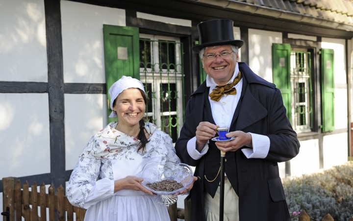 A woman in a period white dress is holding a plate of cookies in her hands. Next to her is an older man in a black man's skirt and top hat. He is holding a teacup in his hand and smiling.