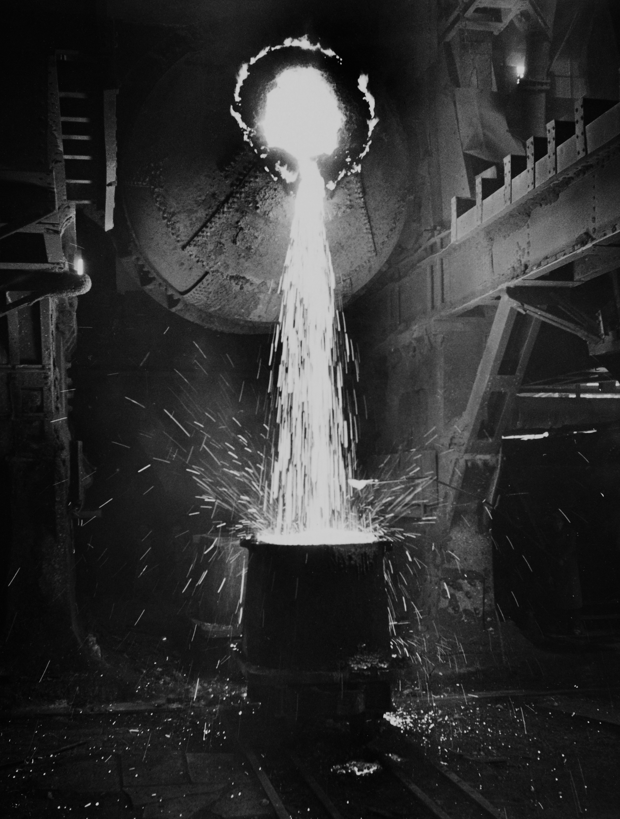 Slag tapping. Black and white photo of liquid flowing down from a round pipe into a barrel. 