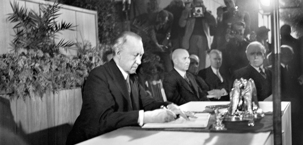 Konrad Adenauer unterschreibt umgeben von der Presse ein Dokument.
