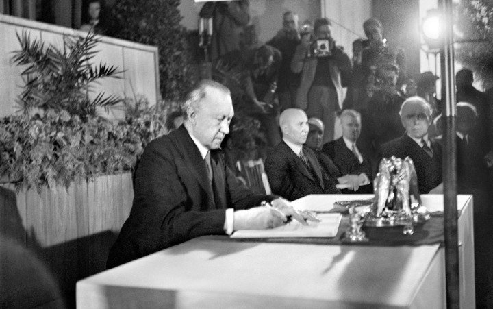 Konrad Adenauer unterschreibt an einem Tisch sitzend und umgeben von vielen anderen Personen ein Dokument