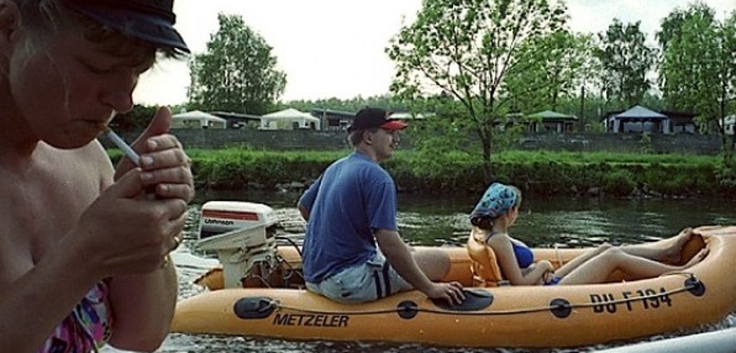 Auf der Ruhr sitzen drei Personen in zwei Schlauchbooten. Im Vordergrund zündet sich eine Frau eine Zigarette an. Im Hintergrund am Flussufer stehen Zelte und Pavillons auf einer Rasenfläche.