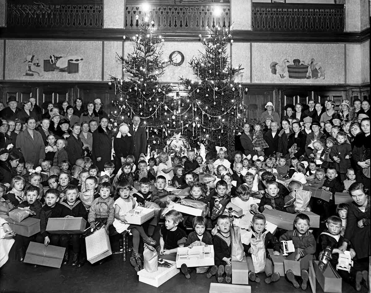 Viele Kinder mit Geschenken sitzen vor zwei großen geschmückten Weihnachtsbäumen. Ältere Kinder und Erwachsene stehen hinter den Kindern neben den Tannenäumen,