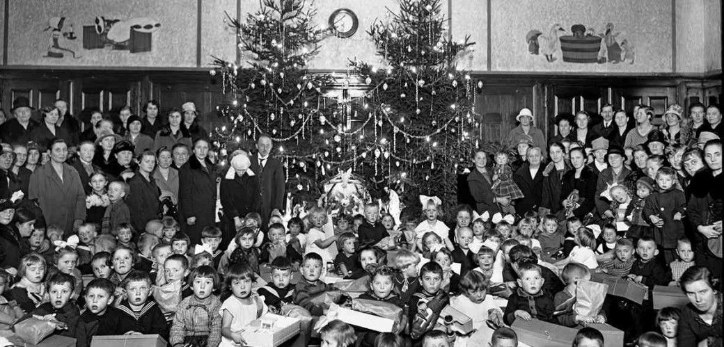 Viele Kinder mit Geschenken sitzen vor zwei großen geschmückten Weihnachtsbäumen. Ältere Kinder und Erwachsene stehen hinter den Kindern neben den Tannenäumen,