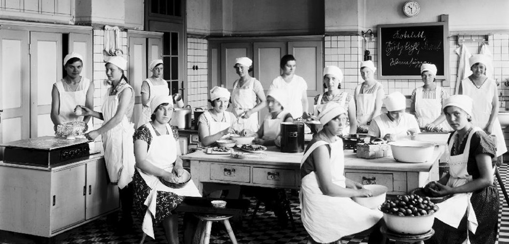 Schwarz-weiß Fotografie einer Gruppe Frauen, die in einer großen Küche kochen und arbeiten.