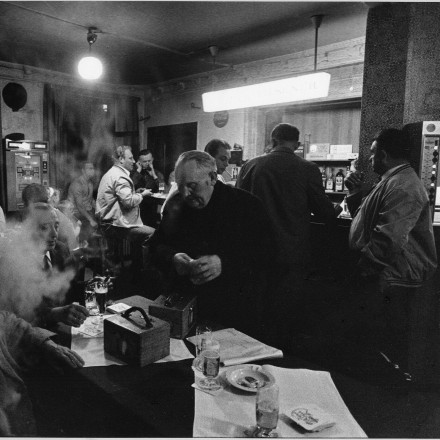Die Schwarzweißfotografie zeigt den Innenraum einer Gaststätte. Mehrere Personen sitzen im Hintergrund am Tresen, im Vordergrund stehen zwei Konstatieruhren auf dem Tisch. Es wird getrunken und geraucht.