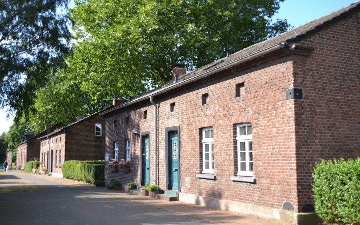 Reihe niedriger Backsteinhäuser mit kleinen Fenstern und Türen, davor Pflanzen und Gartenmöbel, im Hintergrund Bäume und ein Weg.