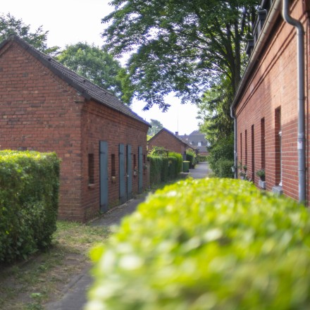 Das Foto zeigt einen Straßenzug in der Siedlung Eisenheim. In der Mitte befindet sich ein ehemaliges Stallgebäude mit grünen Holztüren. Am rechten Bildrand ist die Fassade eines Wohnhauses sichtbar. Beide Gebäude haben Mauern aus roten Backsteinen.