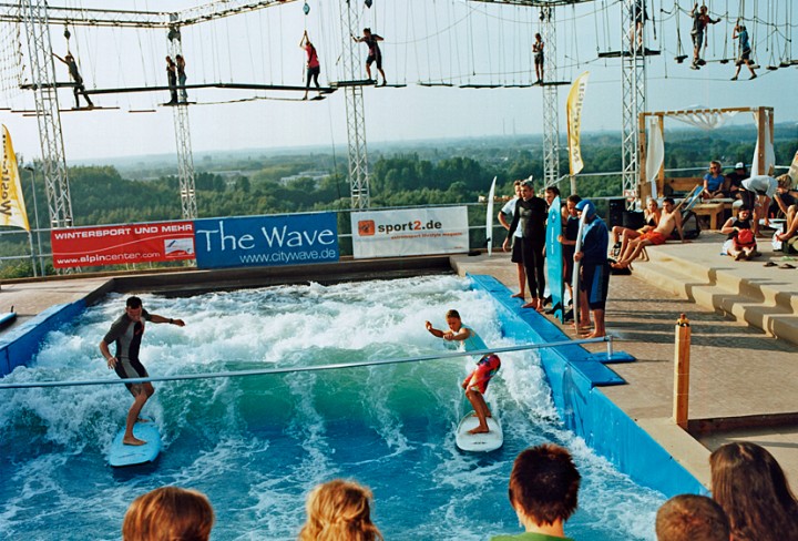 Zwei Surfer auf einem Wellenbecken, dahinter ein Klettergarten, im Vordergrund sitzen Zuschauer