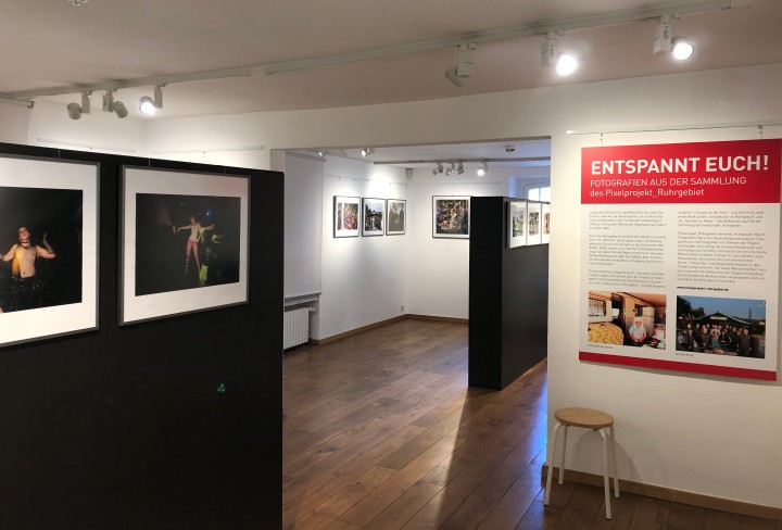 Blick in die Fotografieausstellung. Links im Vordergrund hängen an einer schwarzen Stellwand zwei Fotografien. Rechts im Mittelgrund befindet sich ein ausstellungsteext an einer weißen Wand.