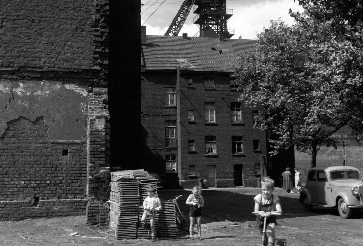 Eine schwarz-weiß Fotografie von einem Jungen auf einem Fahrrad und zwei weiteren spielenden Kindern links von ihm. Im Hintergund ragt der Förderturm der Zeche Westende hinter den Häusern hervor.