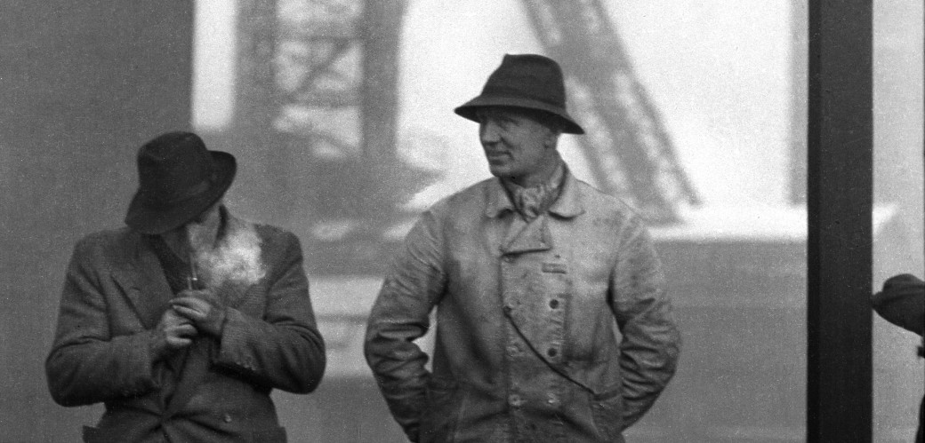 Eine schwarz-weiß Fotografie von zwei Männern mit Hut, die vor dem Förderturm der Zeche Osterfeld stehen. Der linke Mann zündet sich gerade eine Zigarette an.