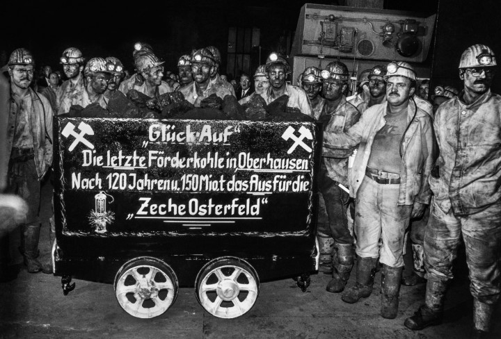 Schwarz-Weiß Fotografie von einer Gruppe Bergleuten, die vor einem beschrifteten Förderwagen posieren. Auf dem Wagen steht: "Glück Auf. Die letzte Förderkohle in Oberhausen. Nach 120 Jahren u. 150 Mio. Tonnen das Aus für die Zeche Osterfeld"