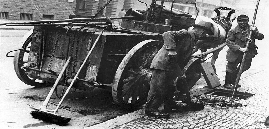 Historische Schwarz-Weiß-Fotografie von zwei Männern, die auf einem Bürgersteig Dreck in eine Kanalöffnung fegen. Hinter ihnen steht ein Pferdekarren