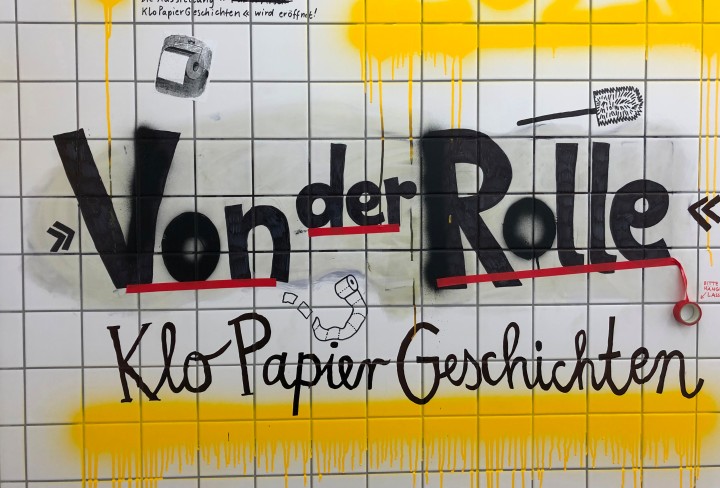 Weiß geflieste Wand mit Graffitti und der Aufschrift "Von der Rolle. KloPapierGeschichten"