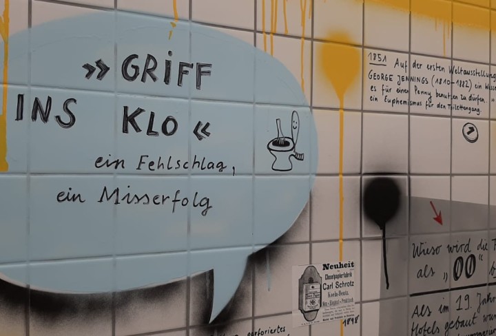 Weiß geflieste Wand mit Beschriftungen und Beschmierungen, im Vordergrund eine blaue Sprechblase: »Griff ins Klo« ein Fehlschlag, ein Misserfolg