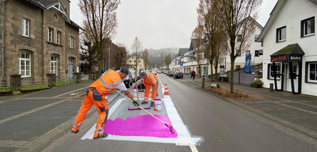 Zwei Männer in orangener Arbeitskleidung verteilen mit einem Besen pinke Farbe auf einer Straße.