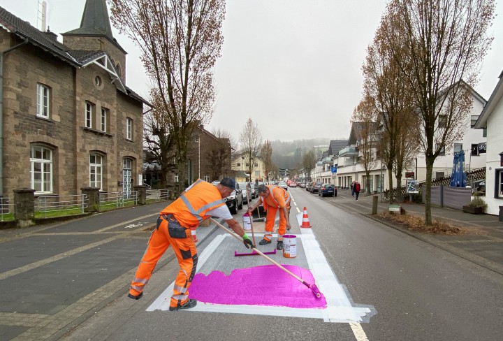 Zwei Straßenarbeiter streichen ein Stück abgeklebte Straße in Pink