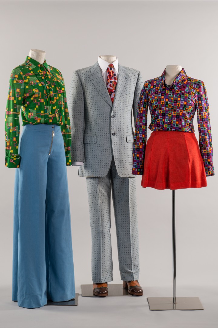 Foto zeigt einen grauen Herrenanzug, ein rot-blaues Kleid und eine blaue Schlaghose mit grüner Bluse für Frauen an Figurinen