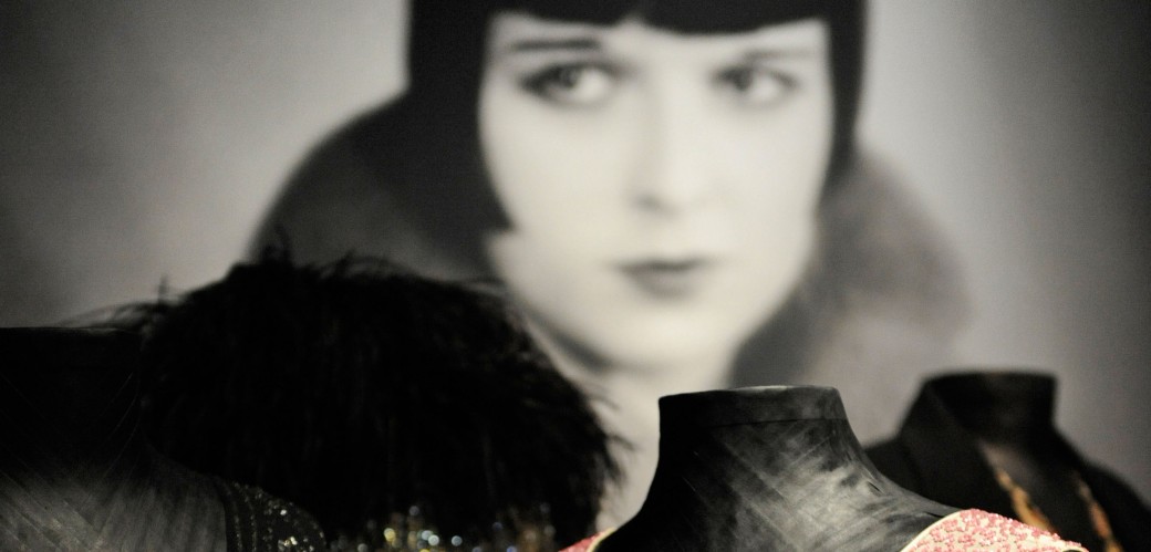 Fotografie eines Frauengesichts in schwarz weiß. Im Vordergrund stehen perlen- und pailettenbesetze Kleider.